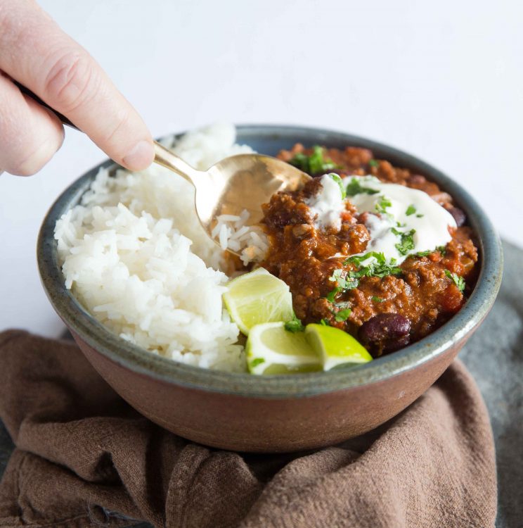 close up shot of hand dunking golden spoon into bowl of chilli, rice and lime wedges