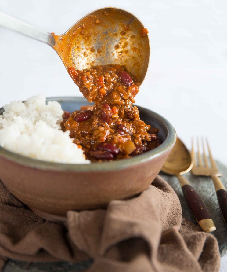 ladle pouring chilli onto rice in small bowl on brown towel next to golden fork and spoon