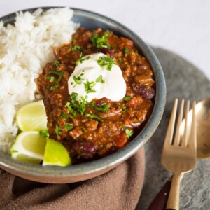 chilli served in small bowl with rice, sour cream and lime wedges next to golden spoon and fork