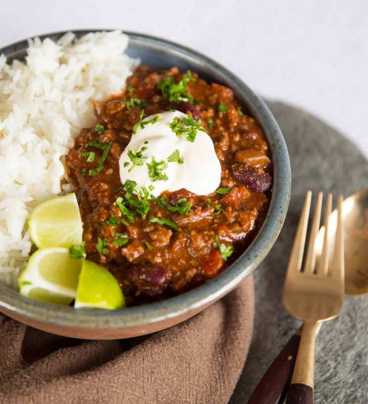 chilli served in small bowl with rice, sour cream and lime wedges next to golden spoon and fork