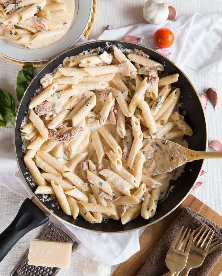 Penne Alfredo with Bacon and Sundried Tomato overhead shot of pasta in skillet surround by forks, garlic, parmesan and basil