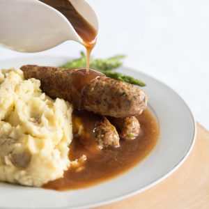 pouring gravy over sausages on white plate with asparagus and mash