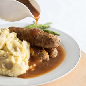 pouring gravy over sausages on white plate with asparagus and mash