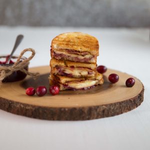 3 grilled cheese stacked on each other on wooden board with glass pot of cranberry sauce by the side