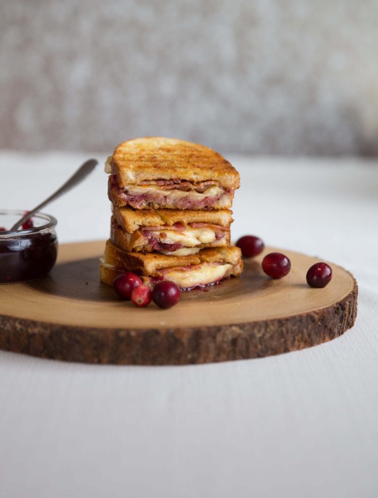 3 grilled cheese slices stacked on each other on wooden board with fresh cranberries scattered around