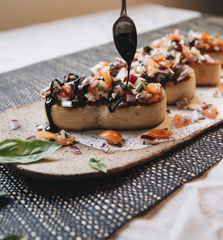 close up shot of teaspoon drizzling balsamic glaze over bruschetta on plate