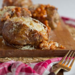 baked potato with beans pouring out on chopping board with 3 more blurred in the background
