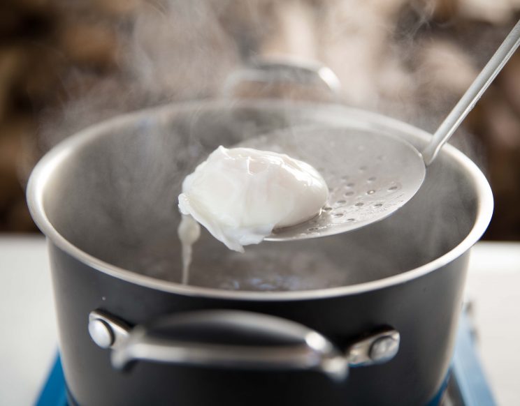 holding poached egg on ladle above pot of steaming water