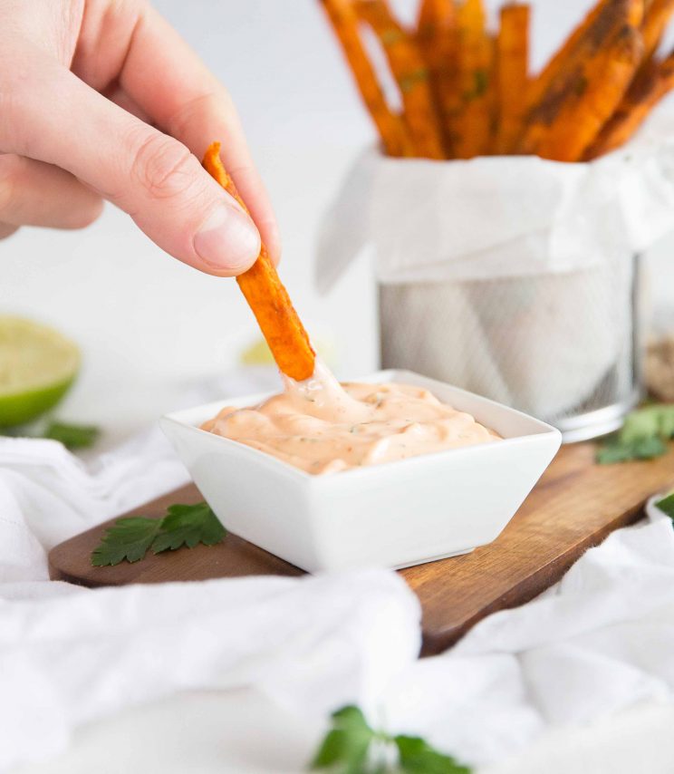 hand dunking single sweet potato fry into small white pot of sriracha mayo