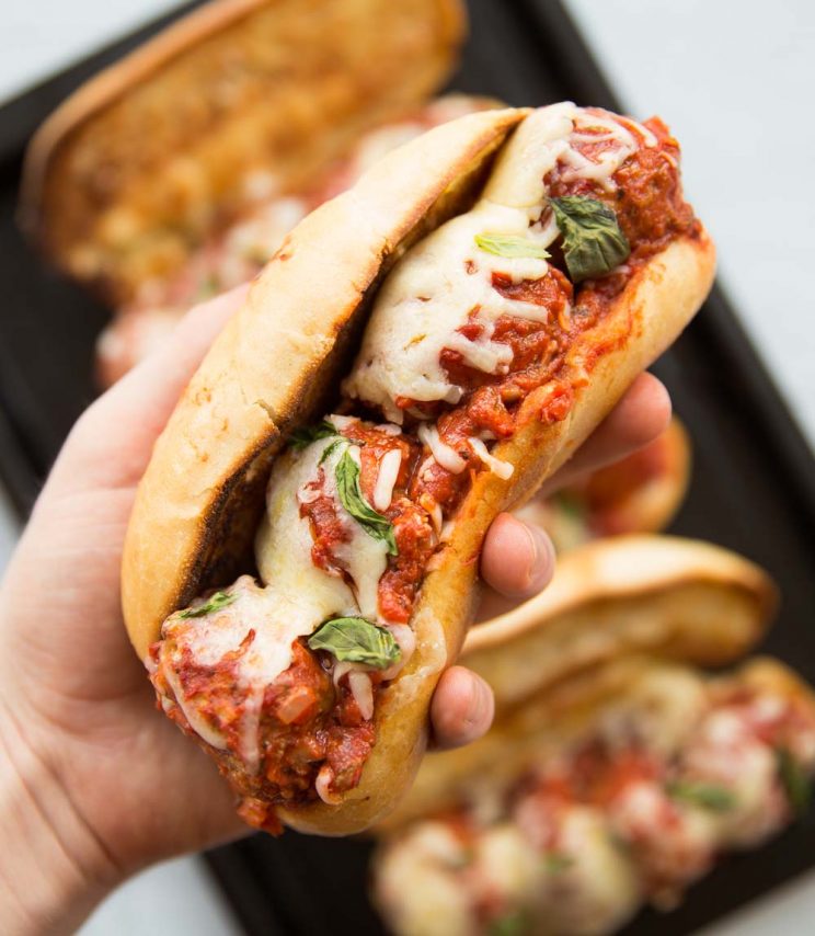 closeup shot of a hand holding meatball sub with melted cheese and 3 subs in the background