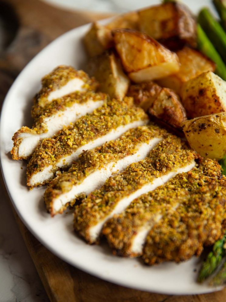 sliced pistachio crusted chicken on small white plate with roast potatoes and asparagus