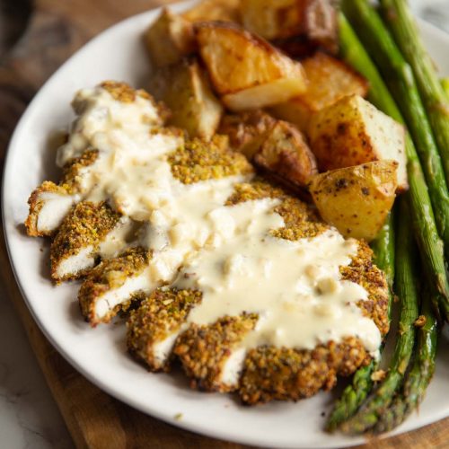 pistachio crusted chicken served on small white plate with potatoes and asparagus with creamy sauce on top