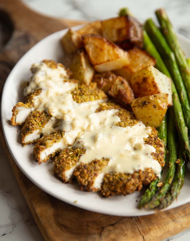 pistachio crusted chicken served on small white plate with potatoes and asparagus with creamy sauce on top