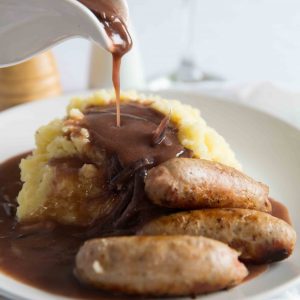 gravy being poured over mashed potato with sausages on small white plate