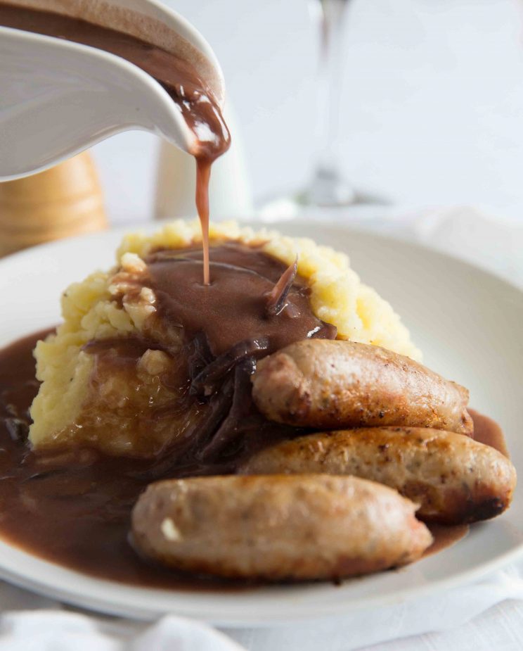gravy being poured over mashed potato with sausages on small white plate