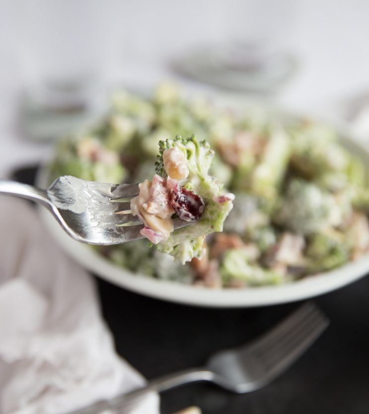 holding fork with broccoli salad on it close up to camera, bowl broccoli salad blurred in background