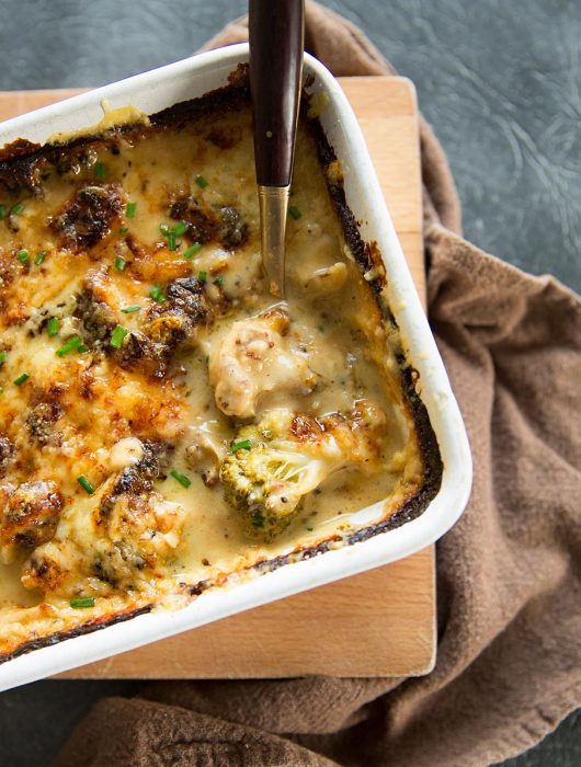 overhead shot of broccoli cauliflower cheese in white baking dish with gold spoon digging in