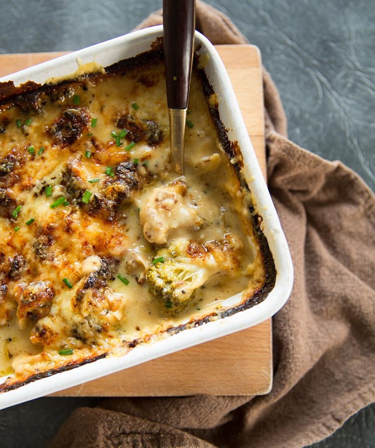 overhead shot of broccoli cauliflower cheese in white baking dish with gold spoon digging in