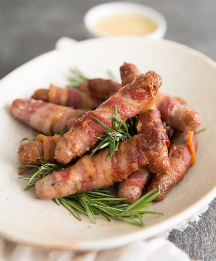 Homemade Pigs in Blankets served with fresh rosemary in a white bowl with dip blurred in background