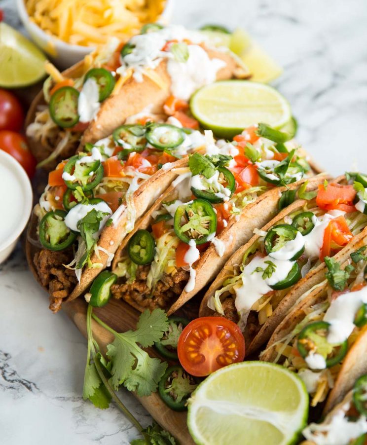 Beef tacos on a wooden board covered in garnishes such as lime, coriander and tomatoes