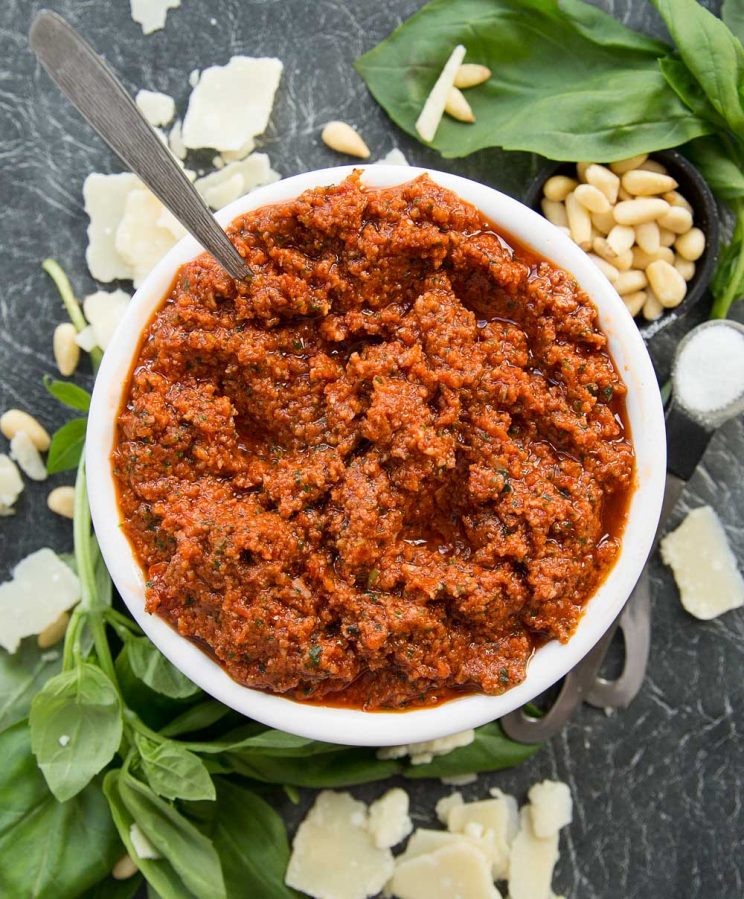 Bowl of Sun Dried Tomato Pesto with spoon poking out surrounded by basil, pine nuts and parmesan