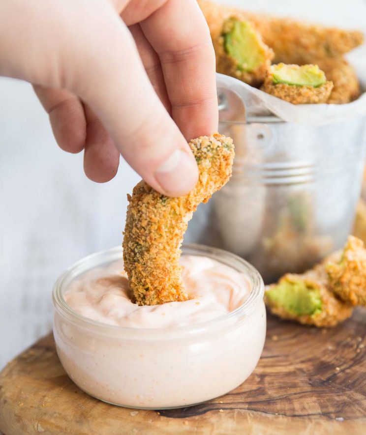 Avocado fry dipping into a pot of sriracha mayo with avocado fries in the background
