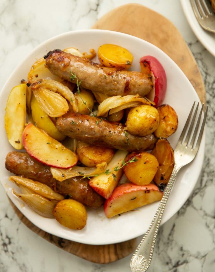 overhead shot of sausage tray bake served on small white plate with silver fork on marble backdrop
