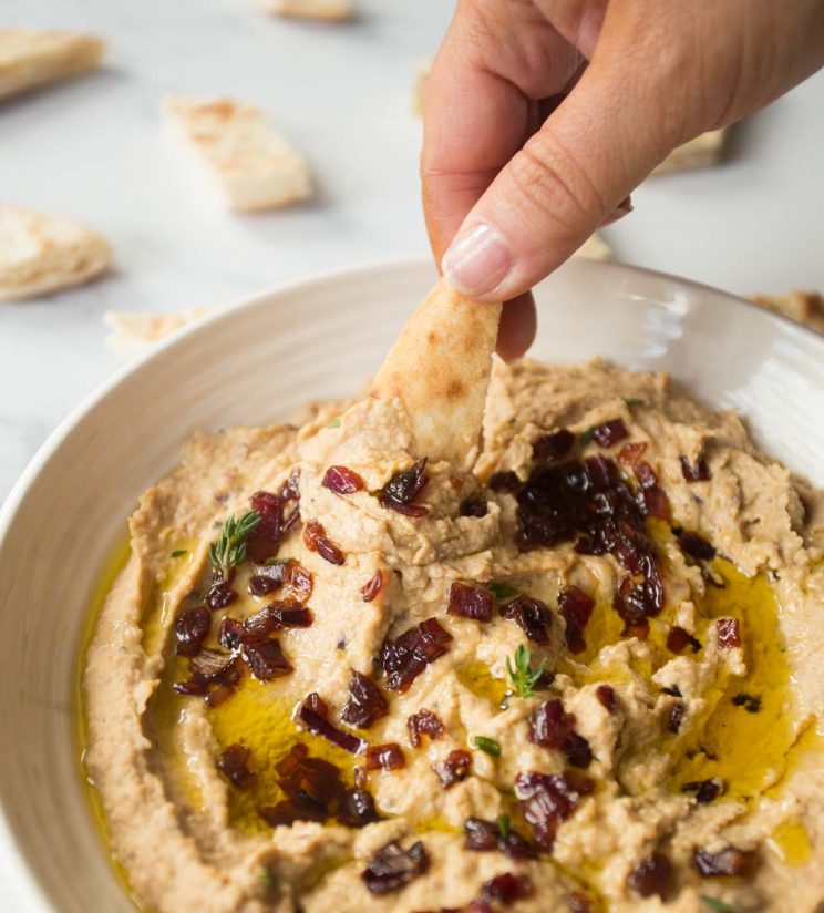 dipping a pita chip into a bowl of hummus garnished with caramelized onions, thyme and extra virgin olive oil