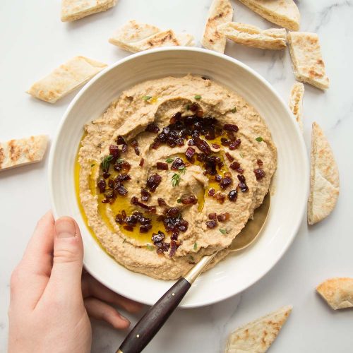 Overhead shot of bowl of caramelized onion hummus with pita bread scattered around and a golden spoon dunked in. Hand holding bowl.