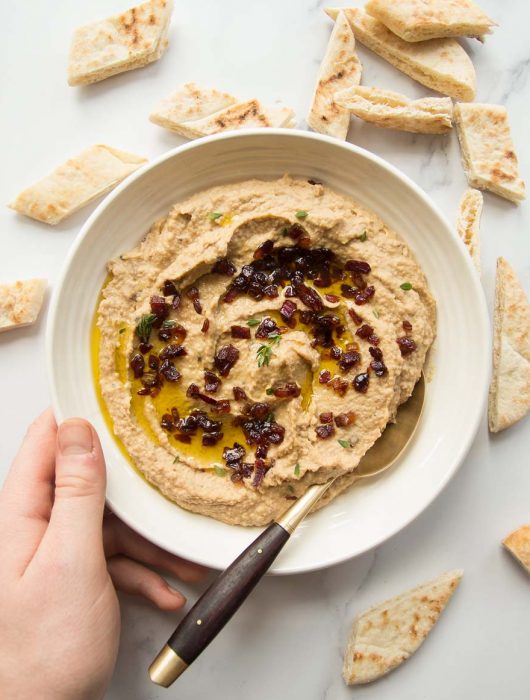 Overhead shot of bowl of caramelized onion hummus with pita bread scattered around and a golden spoon dunked in. Hand holding bowl.