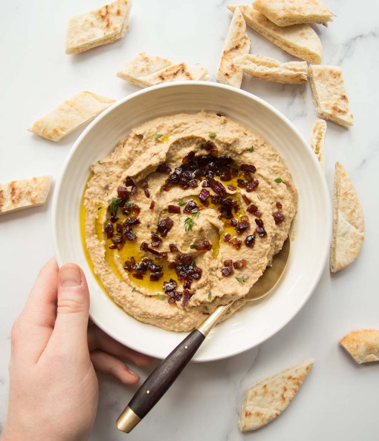 Overhead shot of bowl of caramelized onion hummus with pita bread scattered around and a golden spoon dunked in. Hand holding bowl.