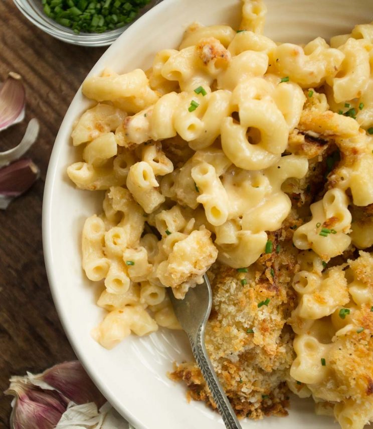 Bowl of roasted garlic mac and cheese with fork digging in and chives and garlic surrounding the bowl