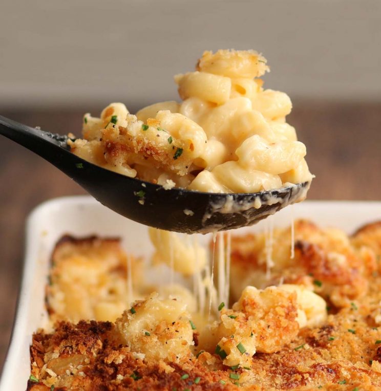 Spoonful of mac and cheese being pulled out of baking dish with cheese dripping down