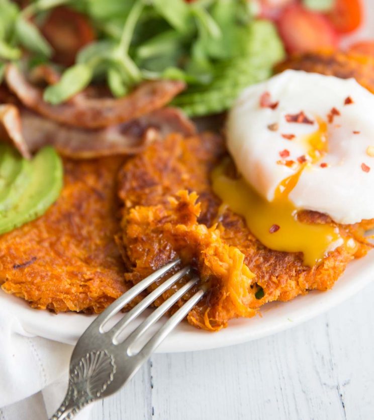 patties on a white plate with a poached egg on top and a side salad in the background. yolk pouring out of the egg on to the hash browns