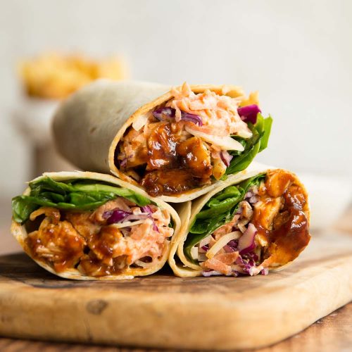 3 bbq chicken wraps stacked on a wooden chopping board with french fries blurred in the background