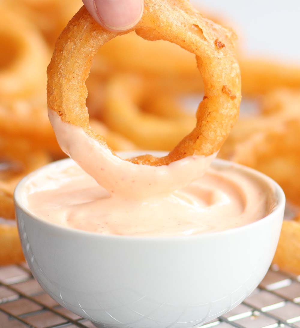 dipping onion ring into srirach mayo with blurred onion rings in the background