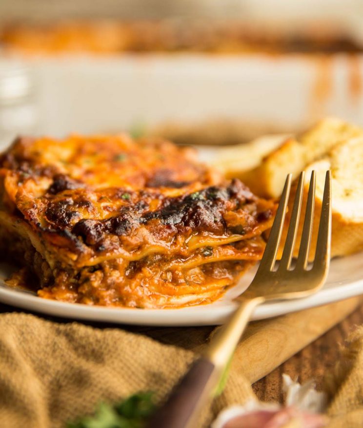 one portion of lasagne served on a plate with baking dish and garlic bread blurred in the background