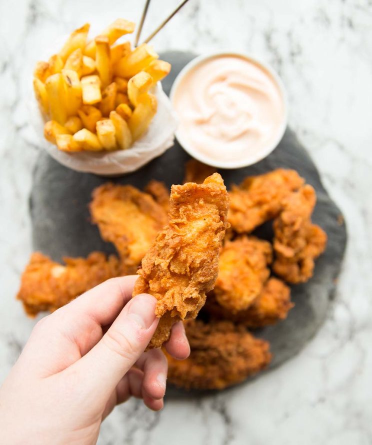 stone slab with chicken strips, chips and dip on top. Holding one chicken strip above the slab with focus on that.