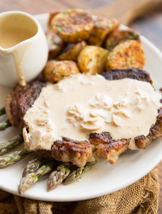 steak on a bed of asparagus with creamy mushroom sauce on top with roast potatoes in the background
