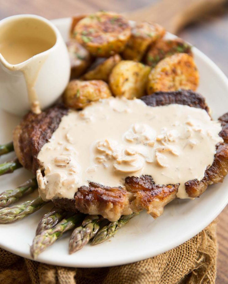 steak on a bed of asparagus with creamy mushroom sauce on top with roast potatoes in the background