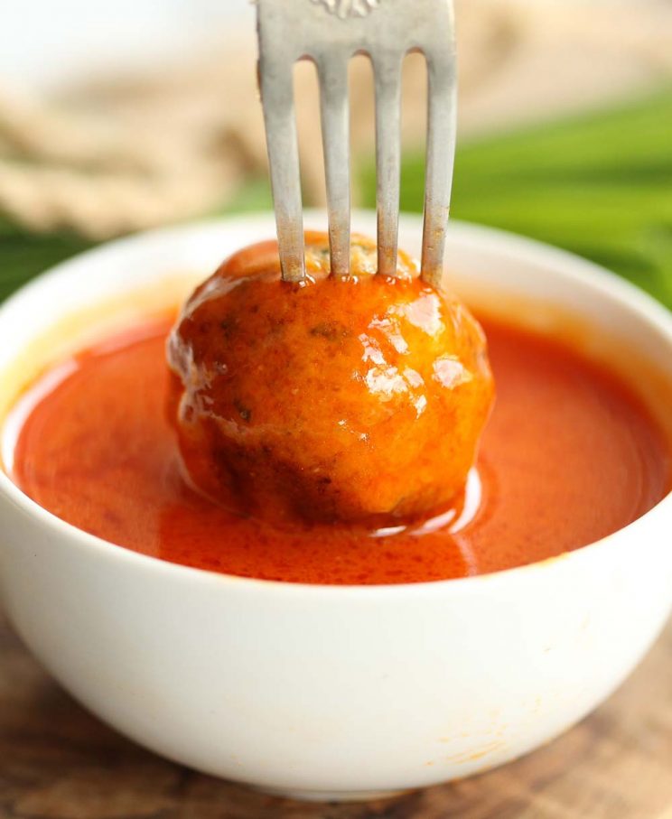 dipping chicken meatball into small pot of buffalo sauce with chives blurred in background