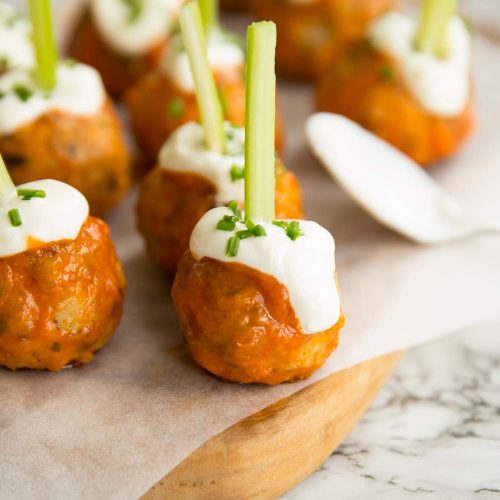 meatballs glazed in buffalo sauce on chopping board with sour cream and chives drizzled on top and celery stuck through the top