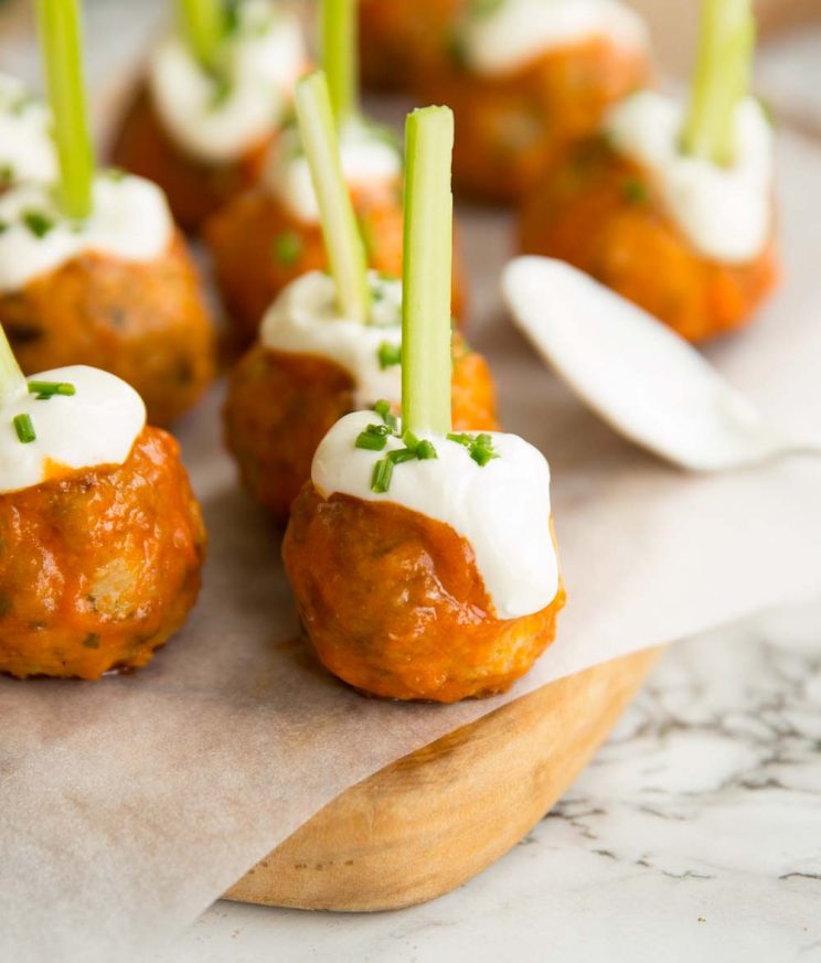 meatballs glazed in buffalo sauce on chopping board with sour cream and chives drizzled on top and celery stuck through the top