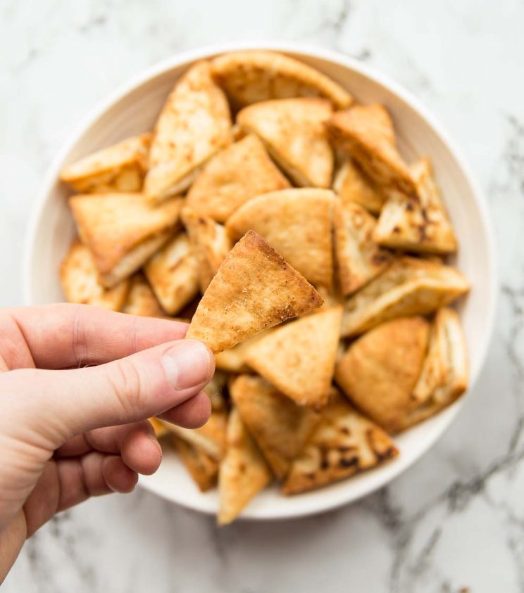 holding a pita chip above a bowl full of pita chips. Focus on holding pita chip.