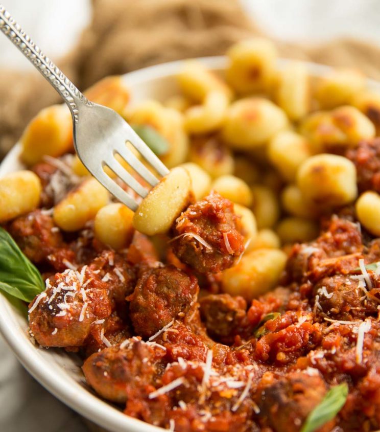 fork holding gnocchi and meatball with a bowl in the background full of gnocchi and meatballs