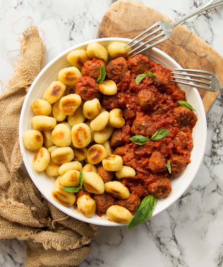 overhead shot of gnocchi and sausage meatballs in white bowl on brown chopping board