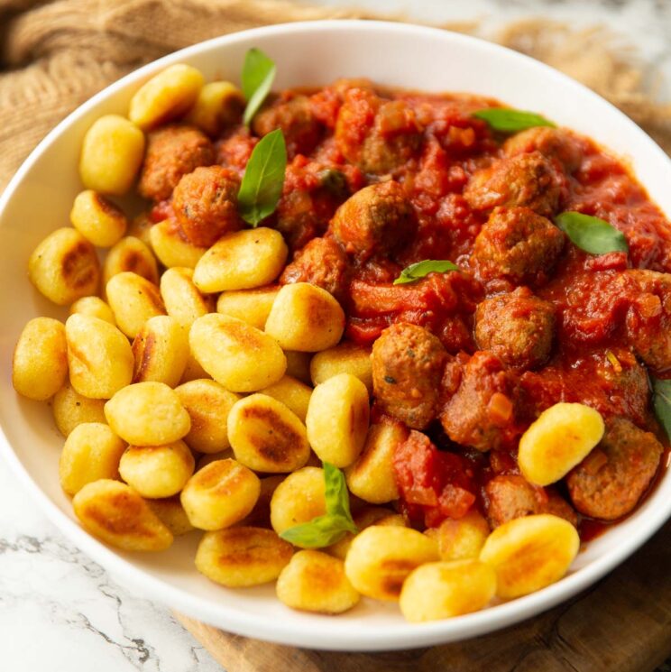 close up shot of toasted gnocchi and sausage meatballs in large white bowl garnished with fresh basil