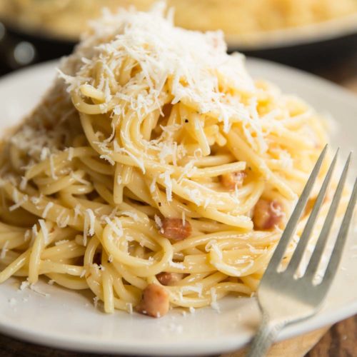 spaghetti carbonara on white plate with saucepan blurred in background and fork resting on plate