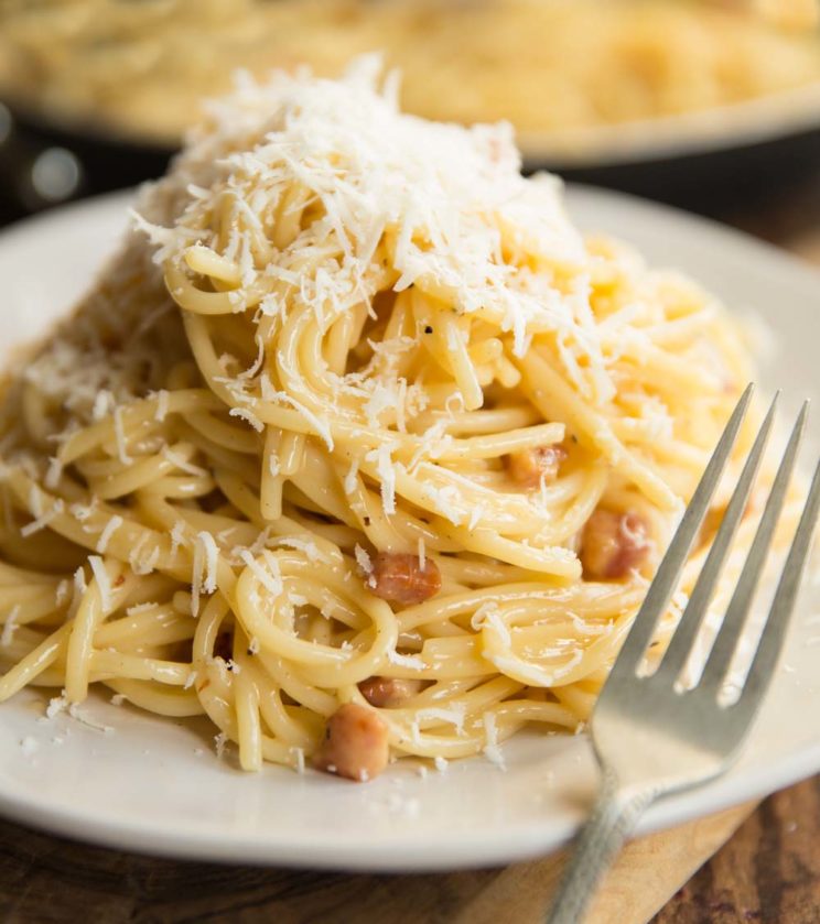 spaghetti carbonara on white plate with saucepan blurred in background and fork resting on plate