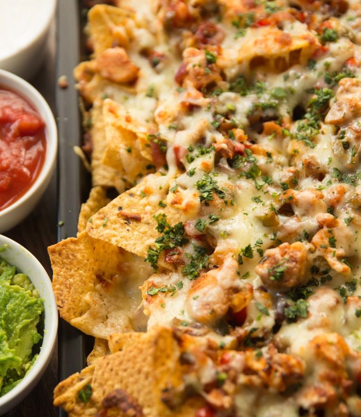 chicken fajita nachos garnished with coriander served on a tray with dips next to them